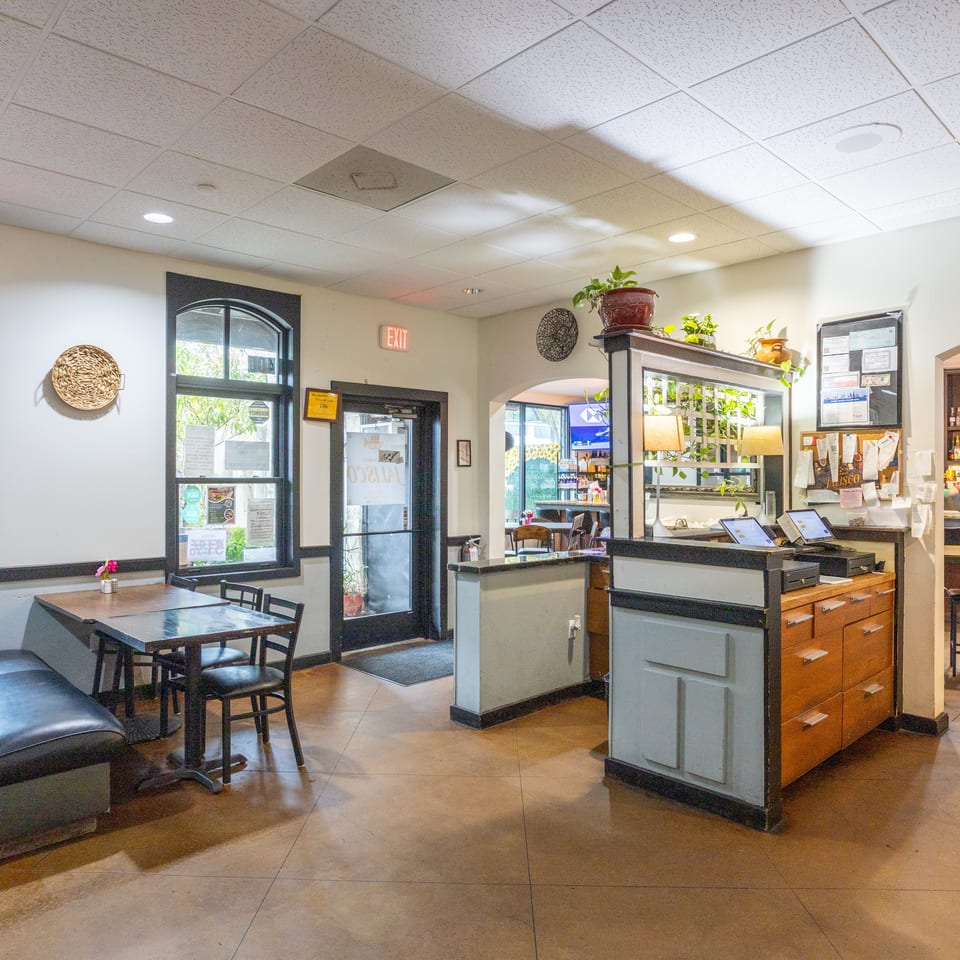 Bright restaurant interior with dining tables, upholstered booths, a host counter, decorative plants, and a stocked bar area.