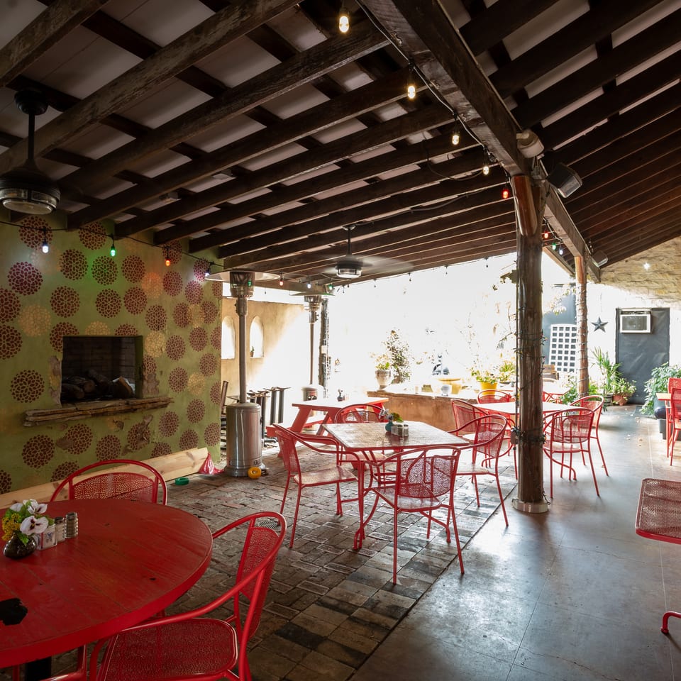 Rustic restaurant patio with red tables and chairs, exposed timber beams, patterned walls, heaters, and a stone fireplace.