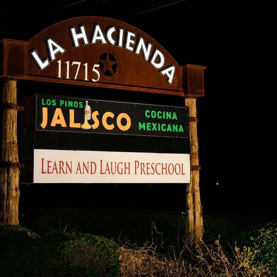 Nighttime wooden sign for La Hacienda 11715, Los Pinos Jalisco Cocina Mexicana, with a Learn and Laugh Preschool sign below.