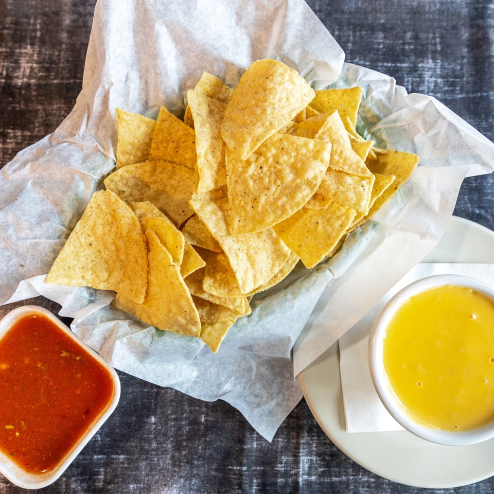 Basket of crispy tortilla chips served with red salsa and yellow cheese dip on a dark tabletop.