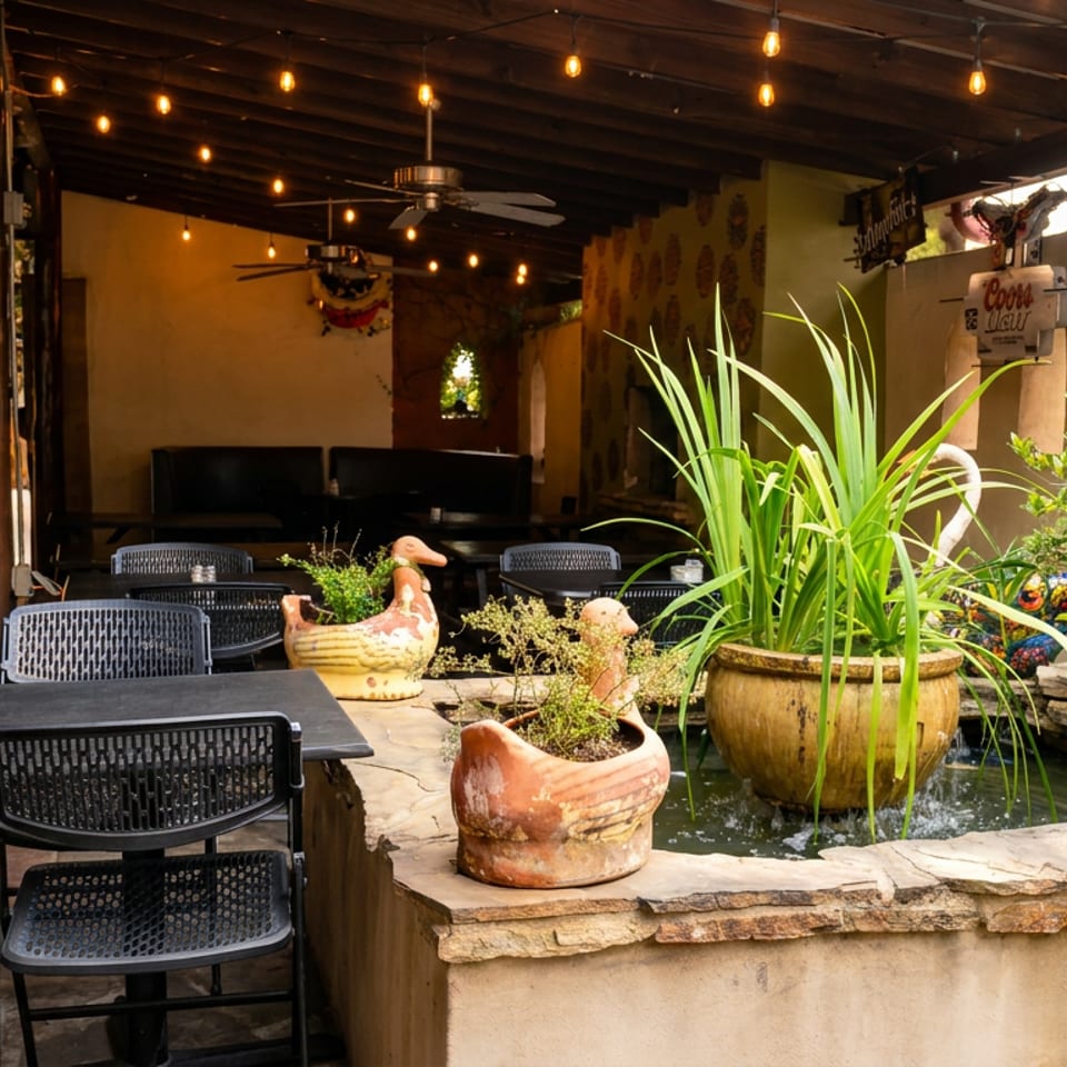 Rustic covered restaurant patio with black tables, string lights, colorful pottery, plants, and a small decorative fountain.