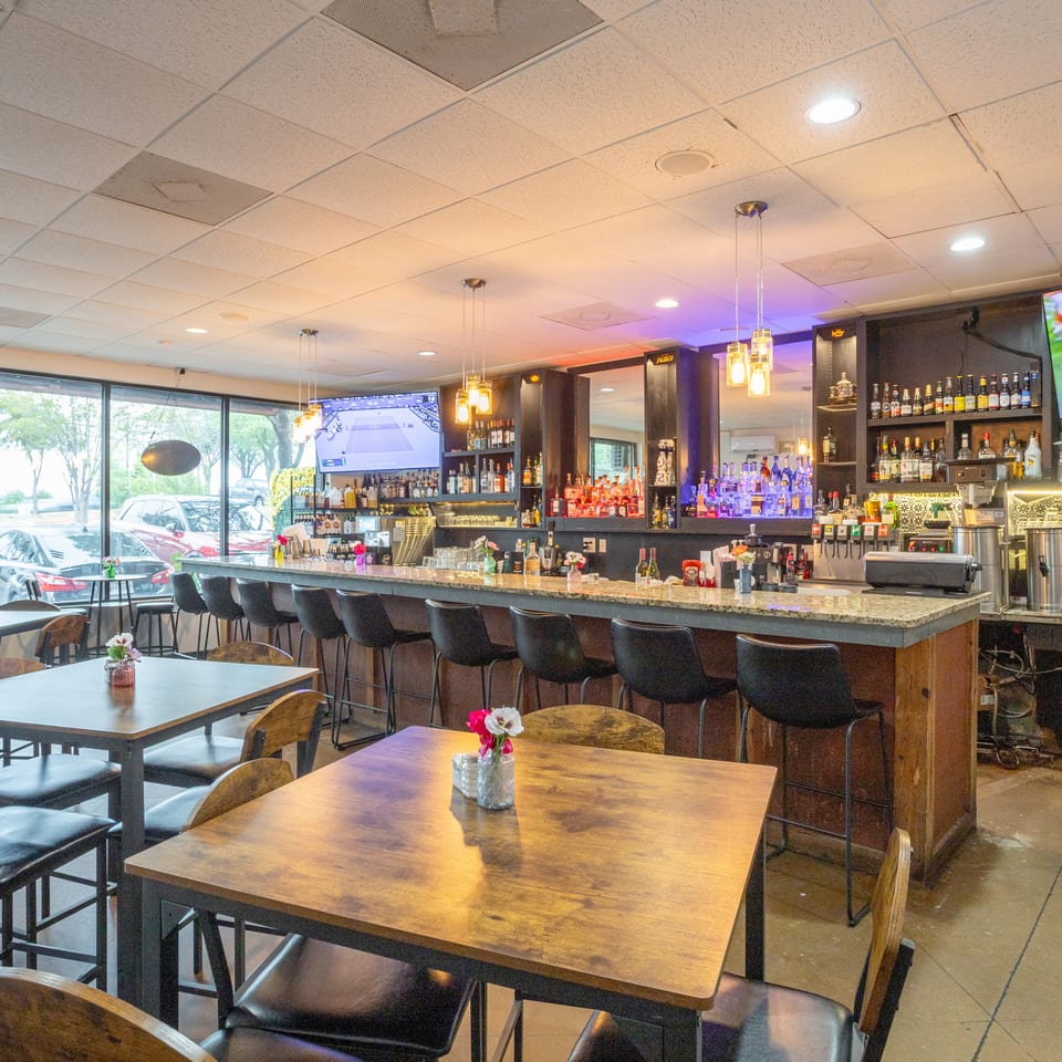 Bright restaurant dining room with wooden tables, black chairs, a stocked bar, hanging lights, and sports playing on televisions.