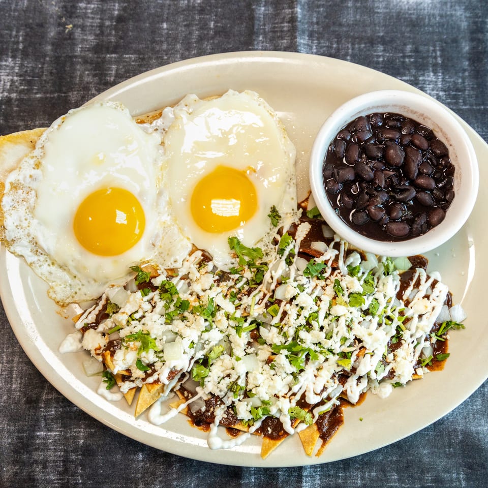 Plate of chilaquiles topped with fried eggs, cheese, onions, and cilantro, served with a bowl of black beans.