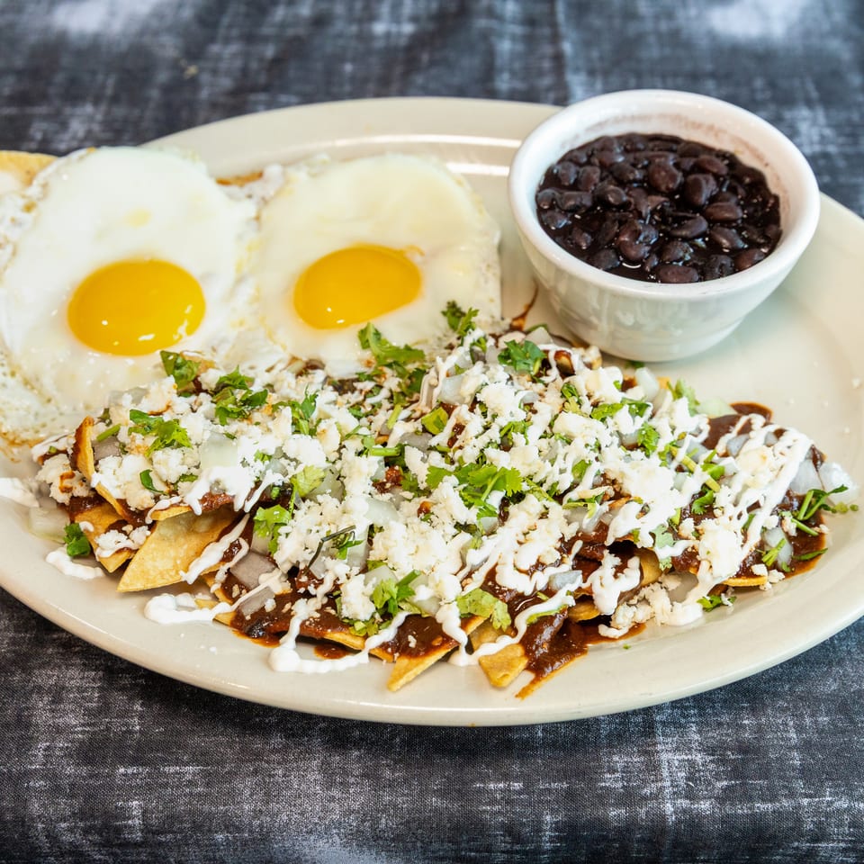 Mexican-style chilaquiles topped with fried eggs, cheese, crema, and cilantro, served with a bowl of black beans.