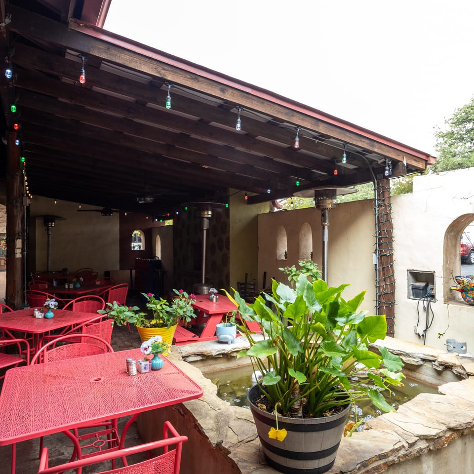 Outdoor restaurant patio with red tables and chairs, colorful string lights, potted plants, heaters, and a small fountain.