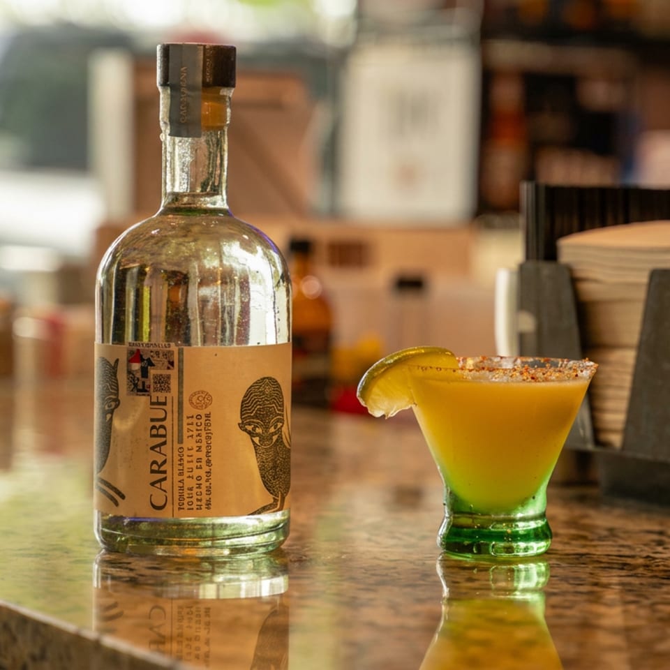 Carabùe tequila bottle and colorful margarita with a salted rim and lime wedge on a sunlit bar.