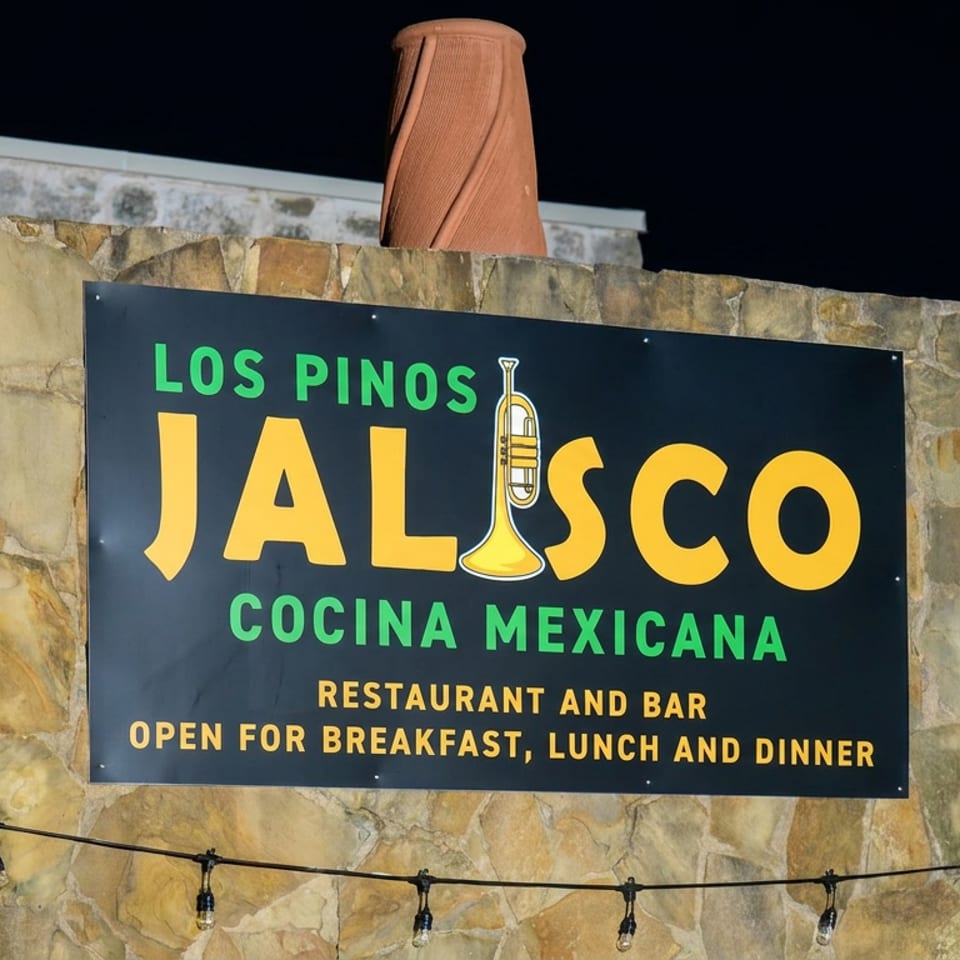Los Pinos Jalisco Cocina Mexicana restaurant sign mounted on a stone facade at night, advertising breakfast, lunch, dinner, and a bar.