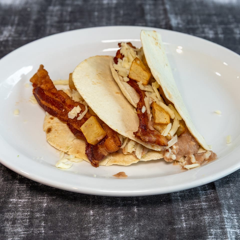 Soft tacos filled with crispy bacon, roasted potatoes, refried beans, and shredded cheese on a white plate.