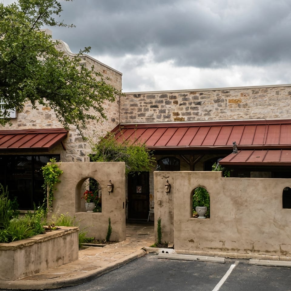 Stone restaurant building with red metal roofs, stucco courtyard walls, mature trees, landscaped planters, and cloudy skies.