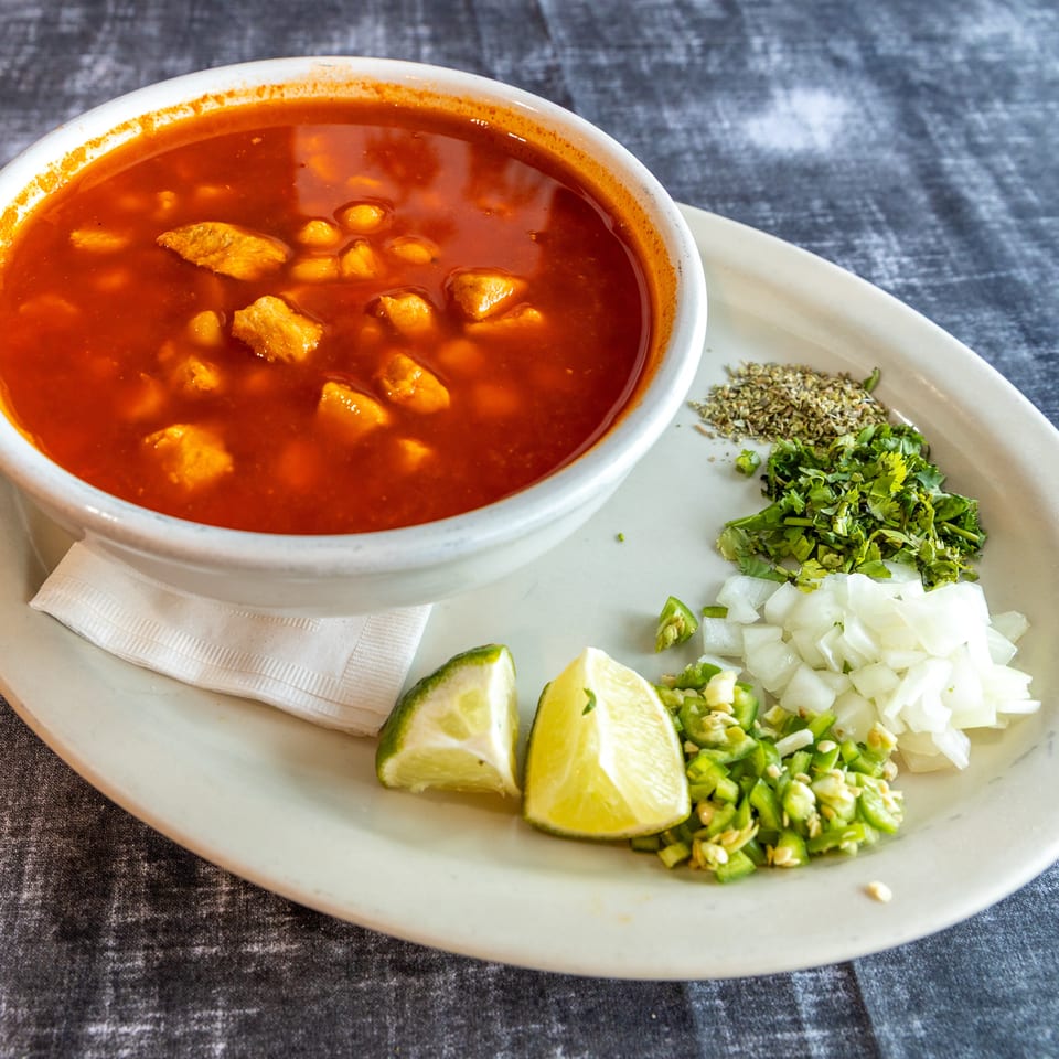 Bowl of red broth with meat and hominy, served with lime wedges, chopped onion, cilantro, jalapeño, and oregano.