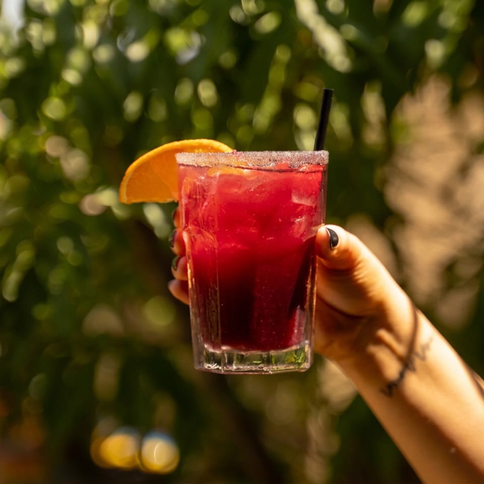 Hand holds a bright red cocktail with a sugared rim, orange wedge, and straw against sunlit greenery.