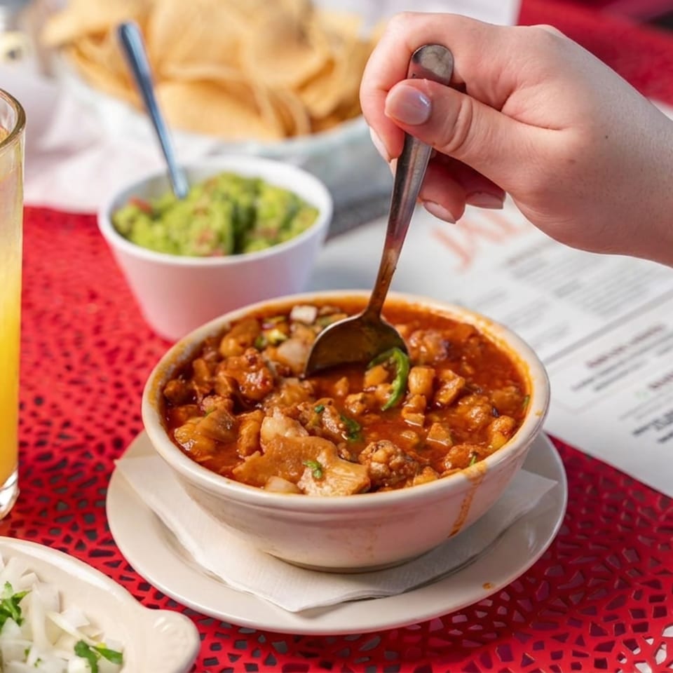 Bowl of chili with beans and toppings, tortilla chips and guacamole, lime rice, and a citrus drink on a red café table.