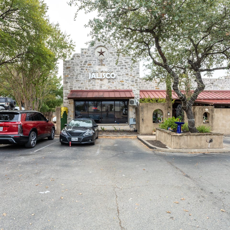 Los Pinos Jalisco restaurant exterior with a stone façade, red awning, star emblem, trees, fountain, and parked cars.