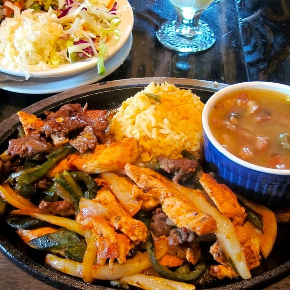 Sizzling skillet with grilled chicken, beef, peppers, onions, rice, beans, and soup, accompanied by a side salad on a restaurant table.