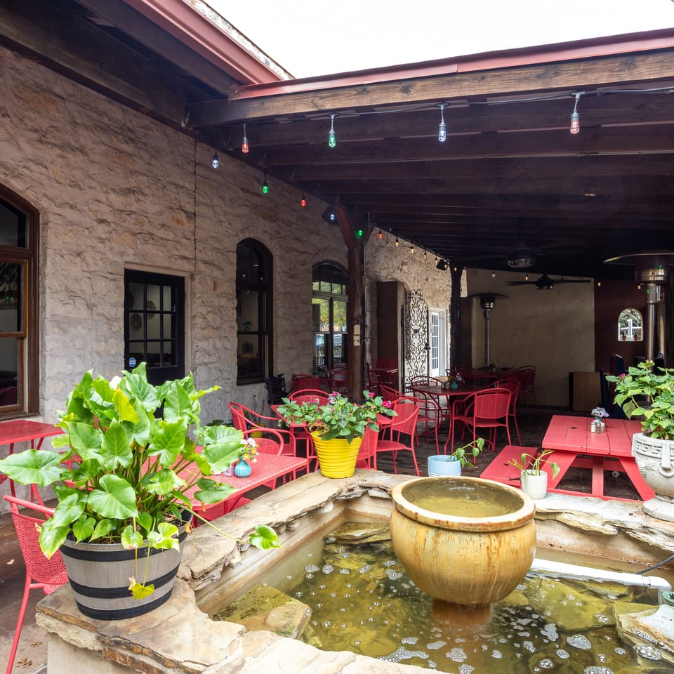 Covered restaurant patio with red tables and chairs, stone walls, colorful string lights, lush plants, and a central fountain.