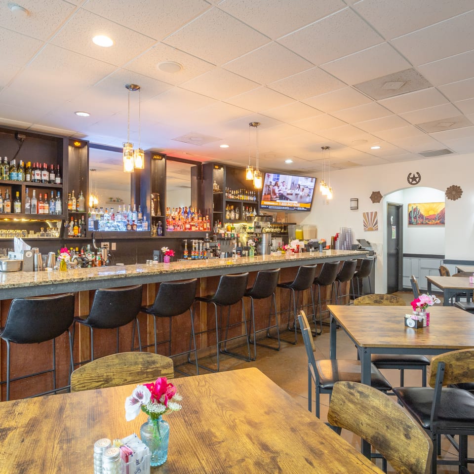 Spacious restaurant interior featuring a fully stocked bar, pendant lighting, black barstools, and wooden dining tables with floral centerpieces.
