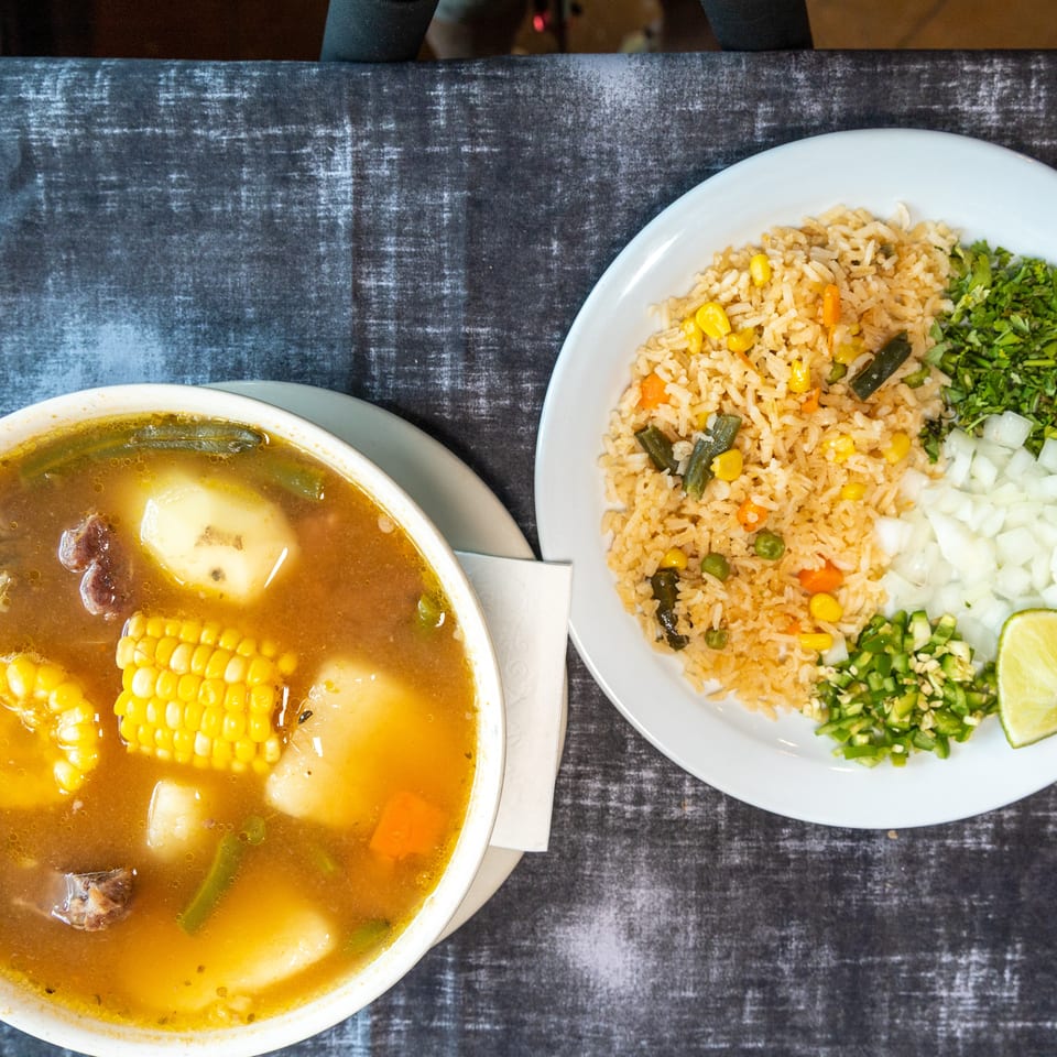 Overhead view of vegetable rice with chopped cilantro, onion, jalapeño, and lime beside a hearty beef and corn soup.