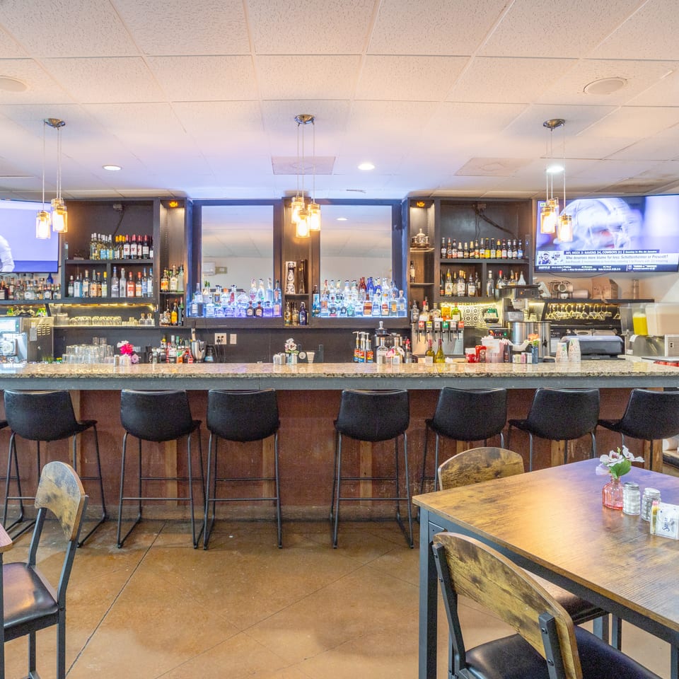 Restaurant bar lined with liquor bottles, pendant lights, television screens, and rows of black stools beside dining tables.